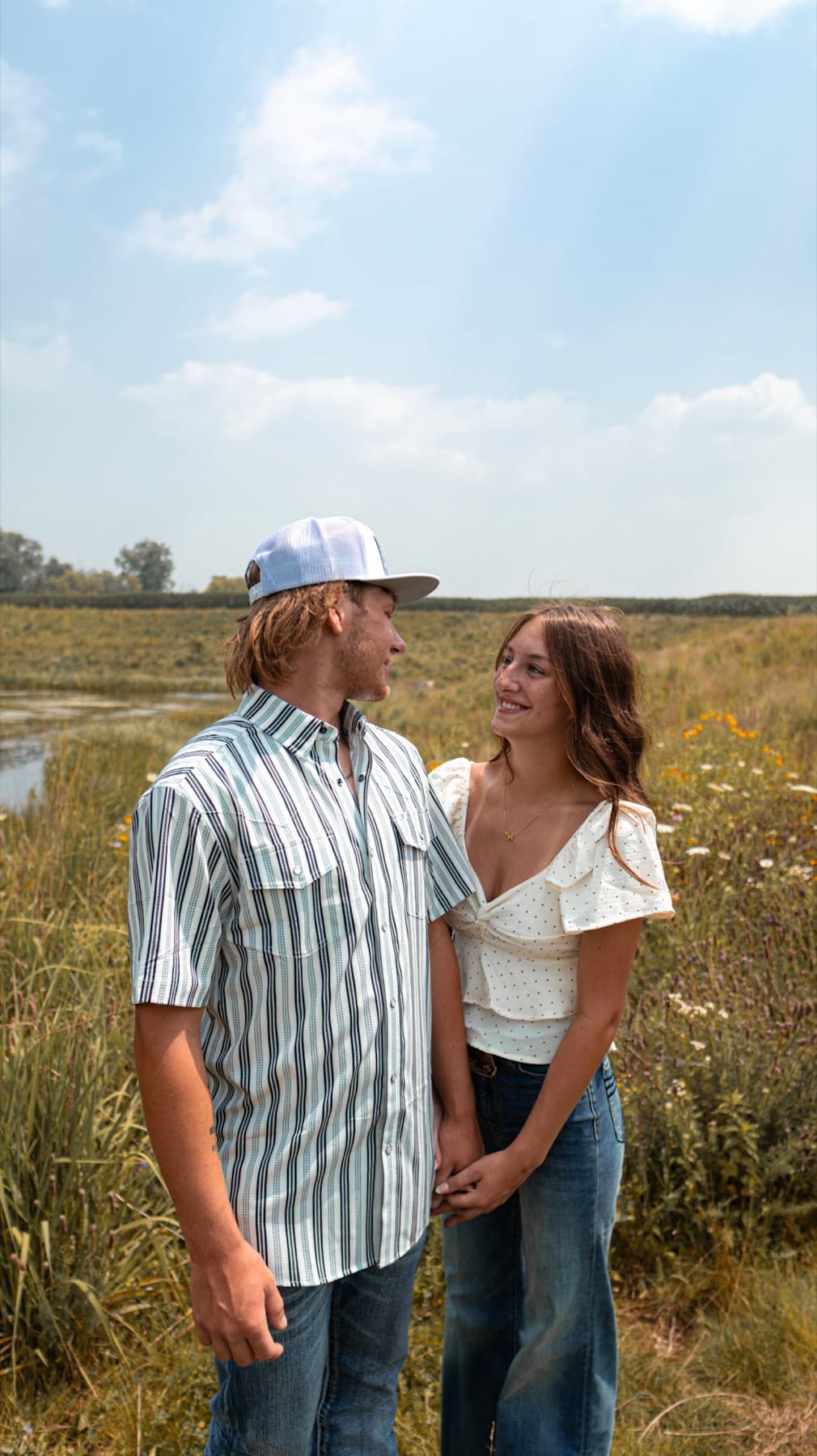 A couple facing each other in a marsh meadow, he in a white cap and striped shirt, she smiling up at him, hands joined.