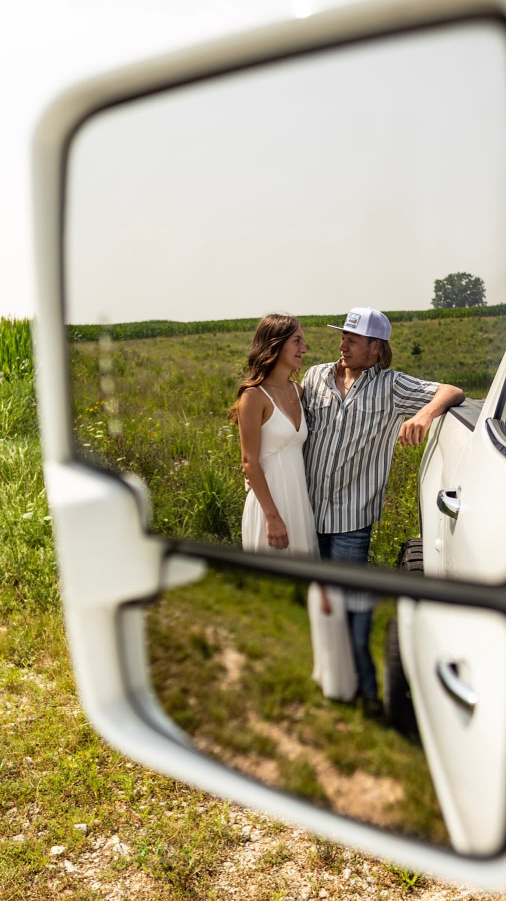 A couple leaning on a white truck in a field, seen in the truck's side mirror.