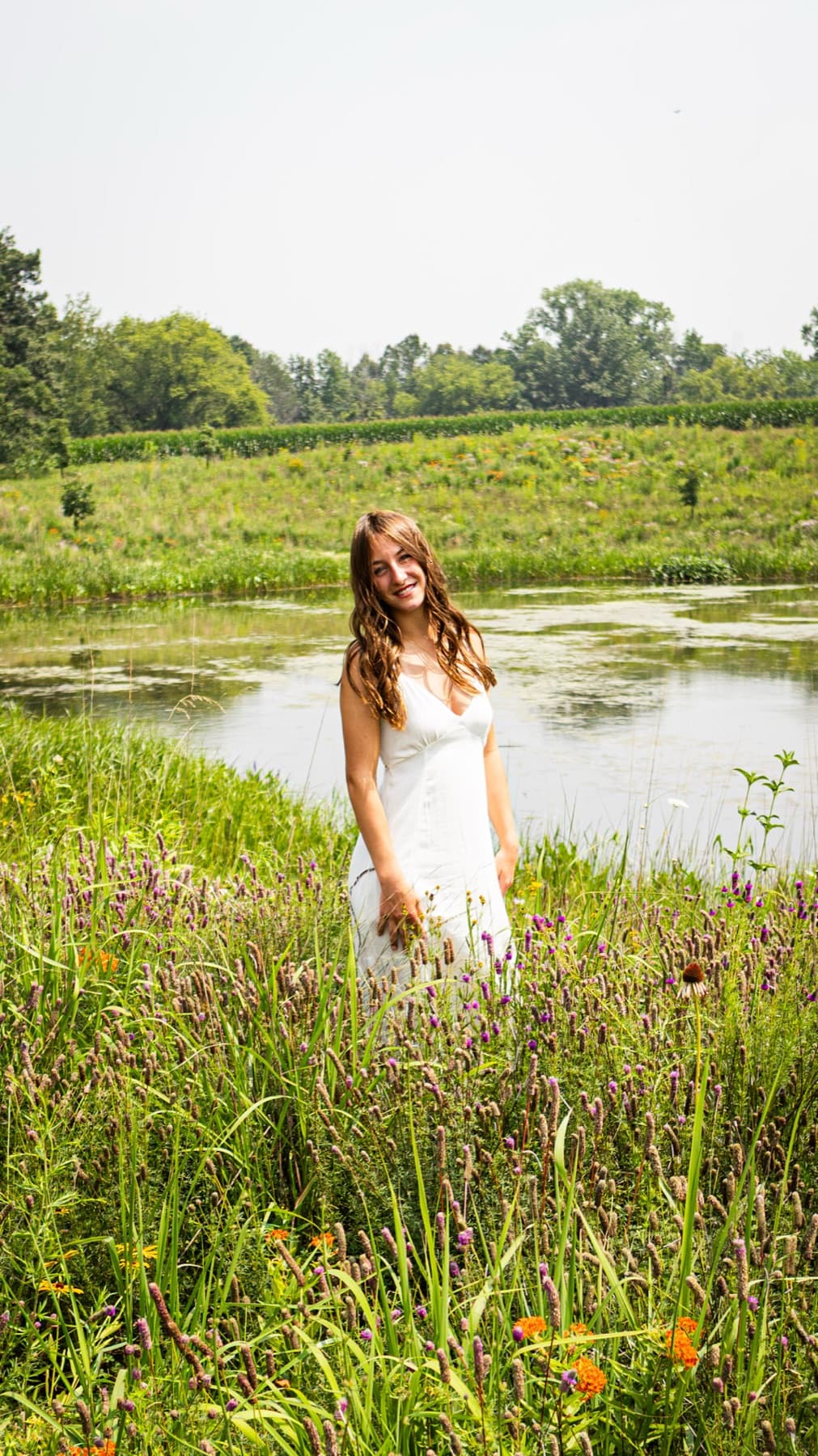 A woman in a white dress standing in tall grass at the edge of a pond, smiling toward the camera.
