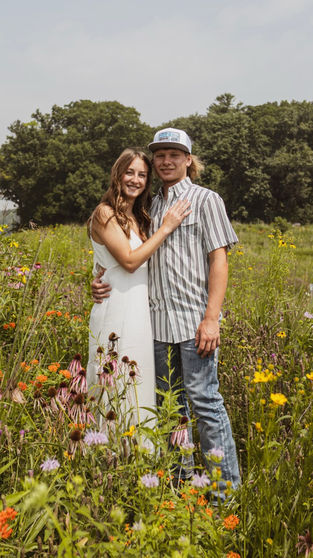 A couple standing close in a wildflower meadow, her arm around his shoulder, oak trees behind them.